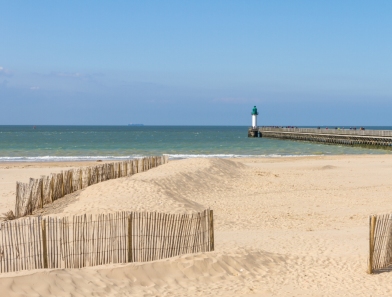 Vue panoramique d’une plage avec dunes protégées, jetée en bois et phare maritime face à l’océan